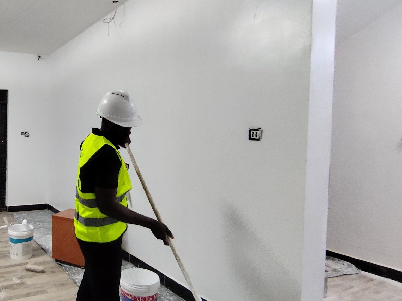 Painter in a hard hat and safety vest rolling white paint onto a living room wall